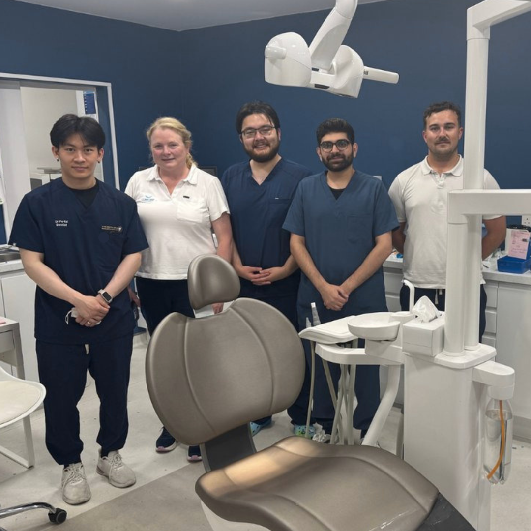 Dental team standing in a modern clinic wearing navy medical scrubs beside a dental chair and equipment.