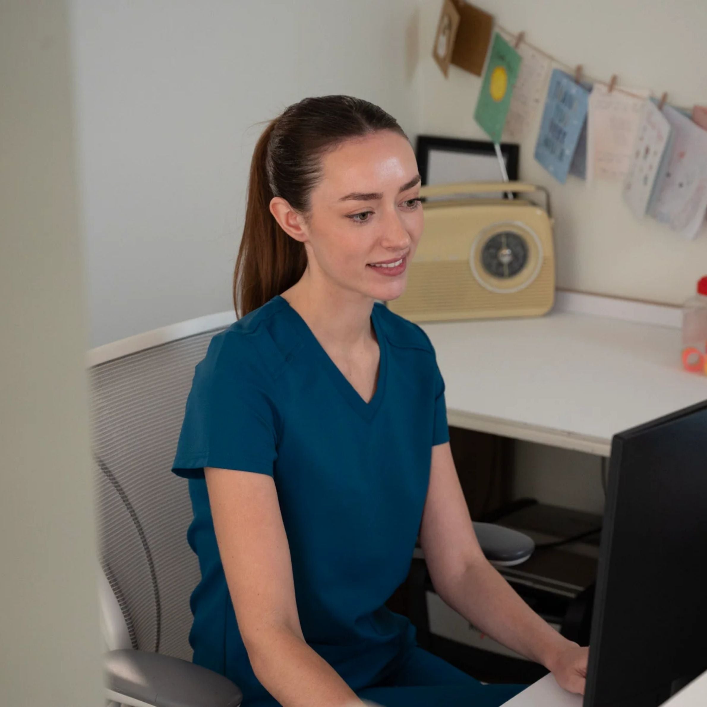 Female HCA wearing caribbean scrubs working at a desk in a clinic reception area, seated at a computer with cards and notes displayed behind her.