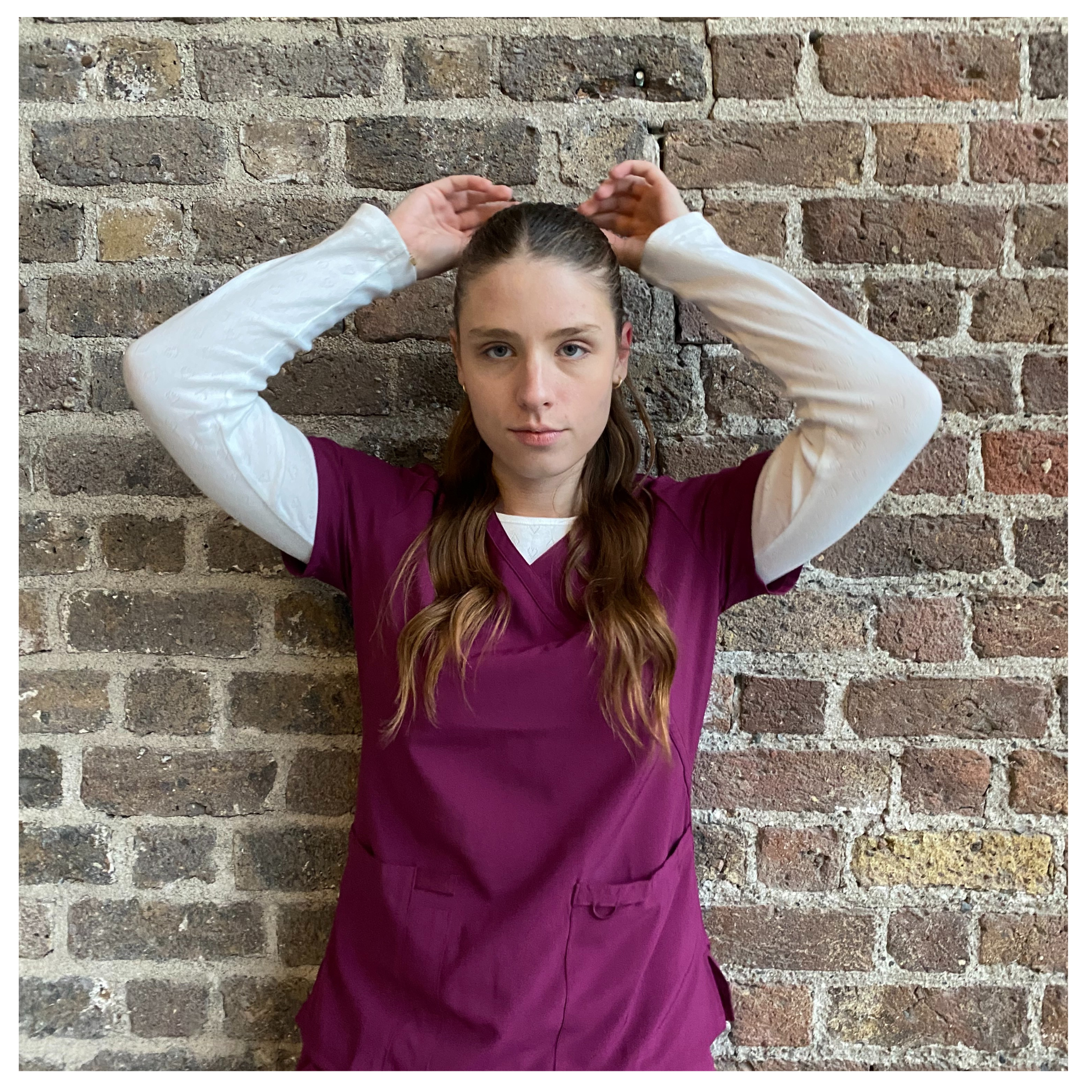 Nurse practitioner wearing burgundy medical scrubs layered over a white long-sleeve top, standing against a brick wall and adjusting her hair, presenting a clean and professional clinical look.