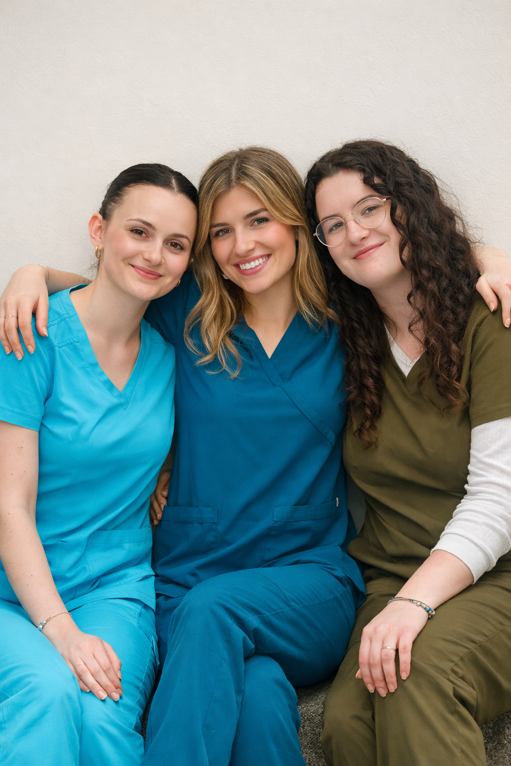 Three female healthcare professionals sitting together and smiling while wearing colourful medical scrubs in teal, turquoise and olive green, representing women working in healthcare.